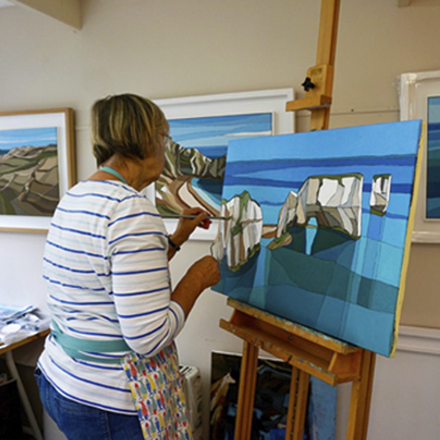 Caz Scott painting at her easel, working on a view of chalk sea stacks in flat blocks of blue, white and green