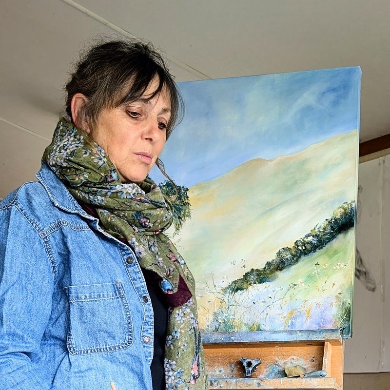 Charlotte Miller in her studio in a denim jacket and scarf, a green and blue landscape on the easel behind her