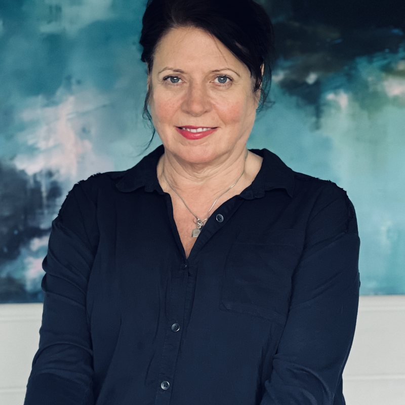 Janet Massey seated in front of one of her own paintings, a pale sea and sky in blue and white