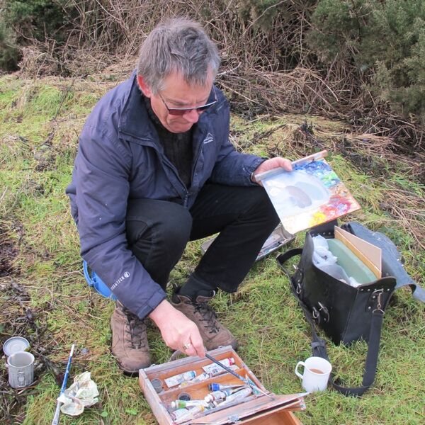 John Boyd crouched on a grass bank painting outdoors, an open paintbox and palette on the ground beside him