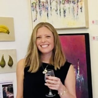 Laura Kedzlie standing among her paintings, a glass in hand