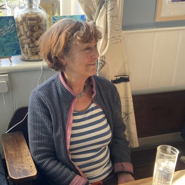 Liz Wright seated in a pub interior, looking off to one side, with her paintings propped on the windowsill behind