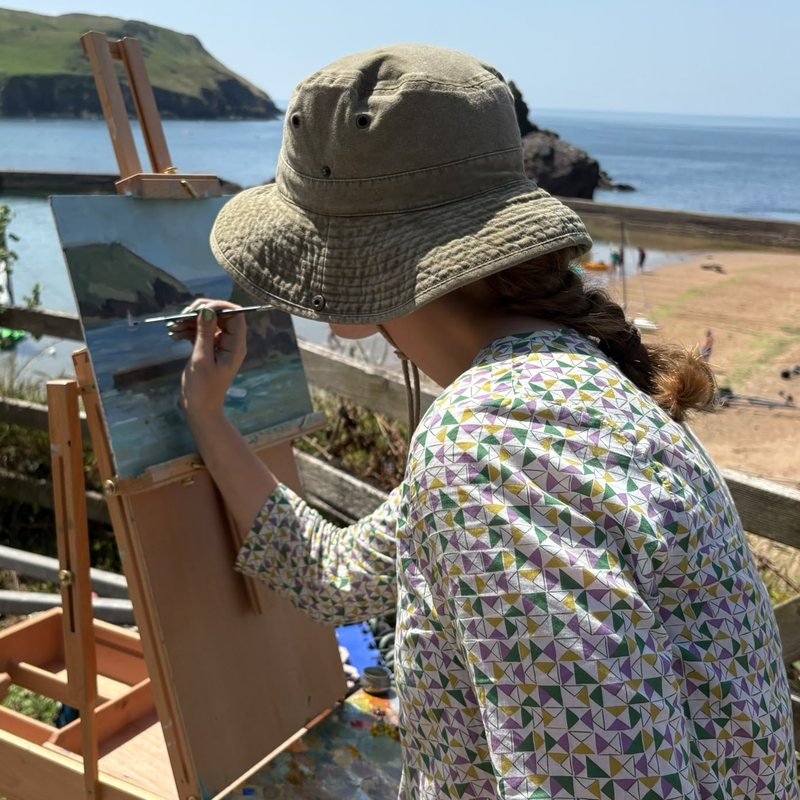 Morgan Willey painting at an easel on the beach in a wide-brimmed hat, a headland and pale sea in front of her