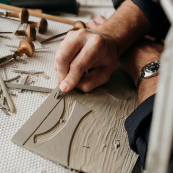 Paul Cleden's hands cutting a lino block with a fine gouge, his cutting tools laid out beside it