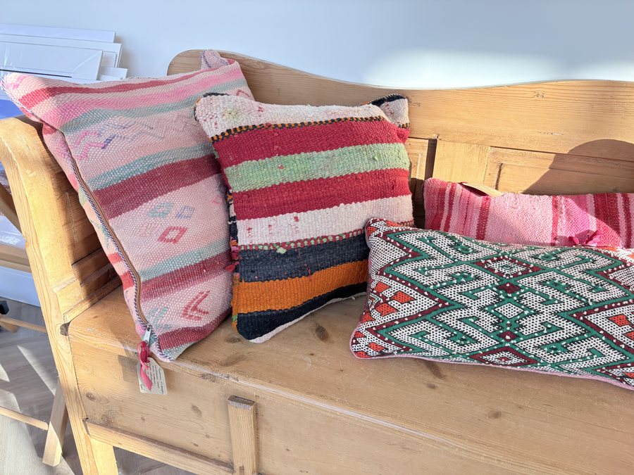 Berber kilim cushions from the Curated Nomad — vintage handwoven wool cushions in pink, red, green and cream stripes and diamonds, ranged along a pine bench in the gallery