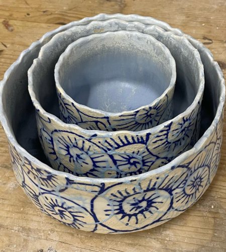 Hand-thrown bowls by Diana Parker — three nested bowls with wavy rims, their outsides carved with concentric blue sgraffito discs on cream, on a bare wooden bench