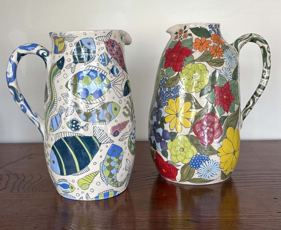 Hand-painted Fish and Flower Jugs by Diana Parker — two hand-thrown porcelain jugs on a dark table, one painted with blue fish, the other with yellow, orange and blue flowers