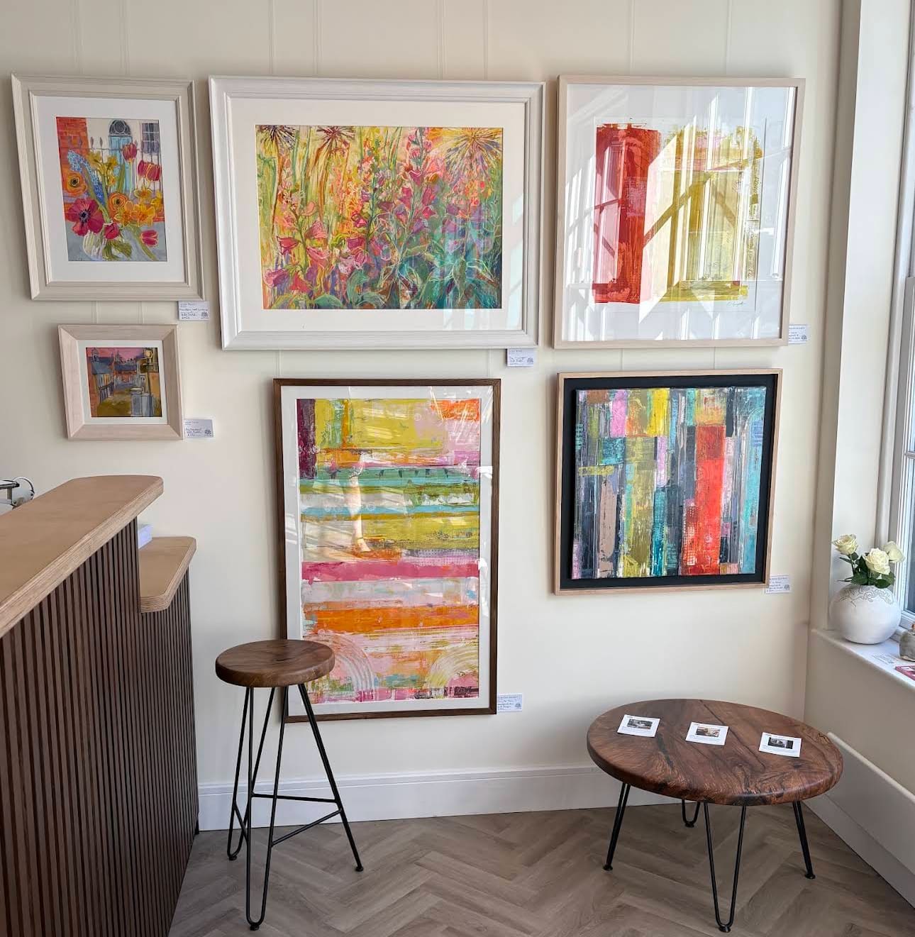A corner of The Walnut Gallery hung with colourful framed paintings and prints, with handcrafted wooden stools and tables