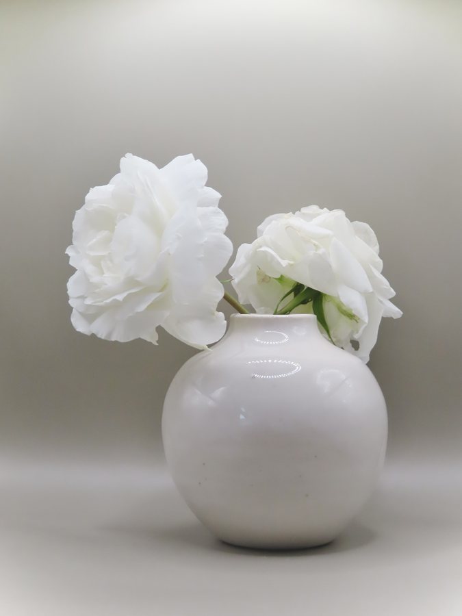 Hand-thrown stoneware vase by Reba Bawden — a round white vase holding two white peonies against a pale grey ground