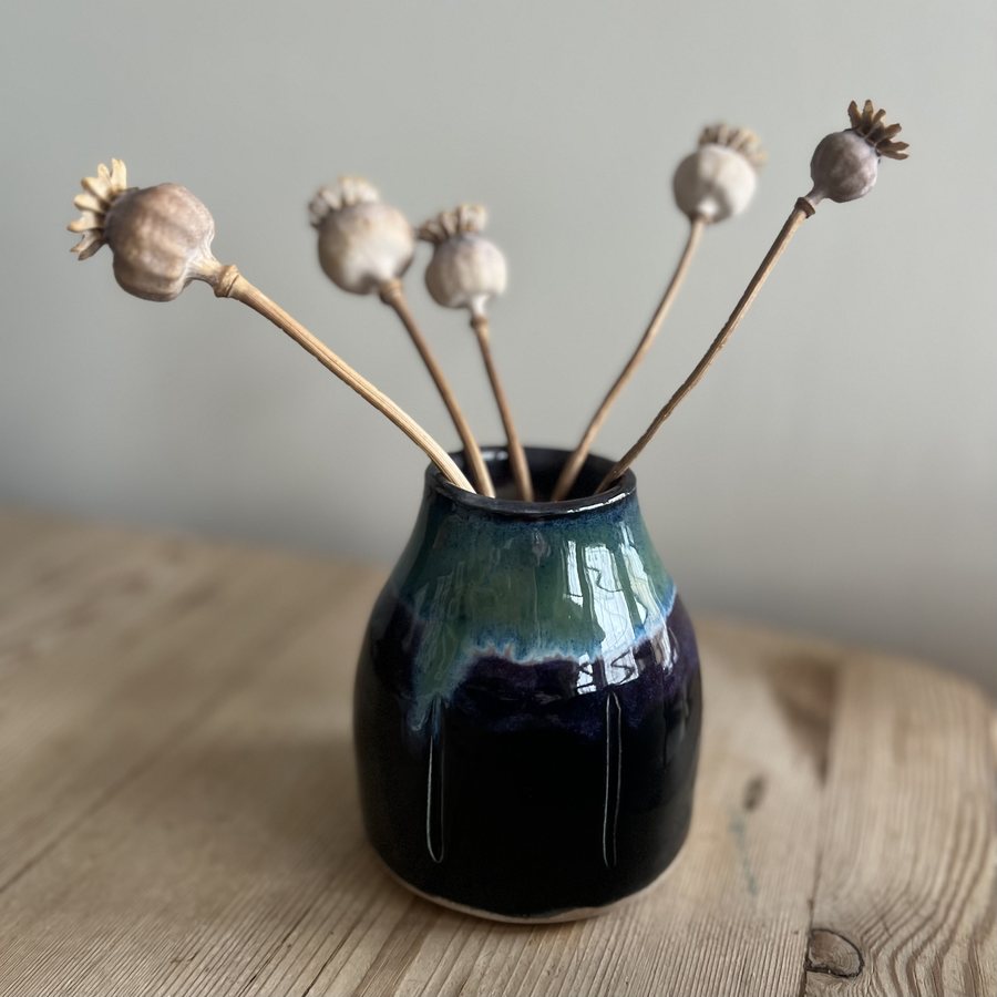 Two Tone Bud Vase by Sharon Oliver — a small hand-thrown vase glazed green over deep blue-black, holding five dried poppy seed heads, on a pale wooden table