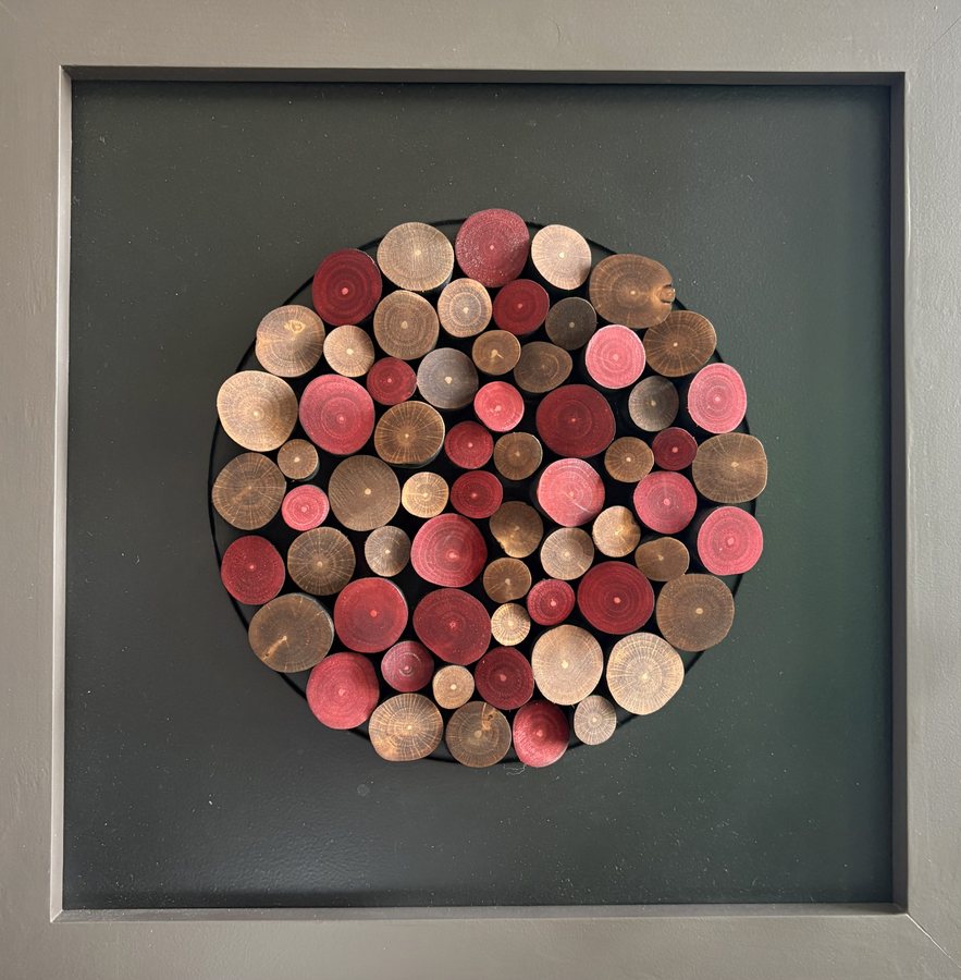 Burst of Hazel — framed wood art by Victor Krause, a disc of end-cut hazel rounds in pink, red and pale wood tones mounted on a grey-green ground